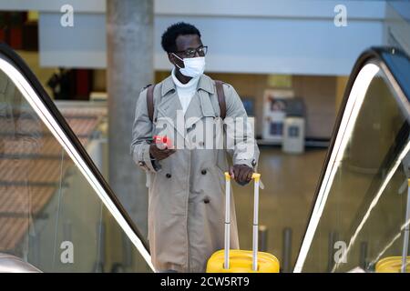 Afro-amerikanischen Reisenden Mann mit gelben Koffer steht auf Rolltreppe in Flughafen-Terminal, tragen Gesicht medizinische Maske, um sich vor Kontakt mit zu schützen Stockfoto