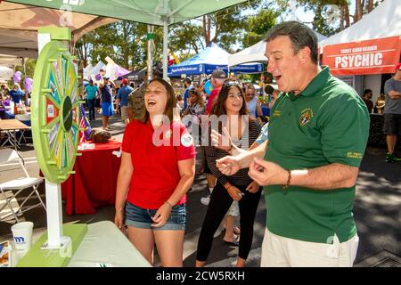 Ein katholischer Diakon gesellt sich mit jungen Gemeindemitgliedern an einem Rad des Zufalls während einer überdachten Straßenmesse Outreach-Ausstellung in Laguna Niguel, CA. Stockfoto