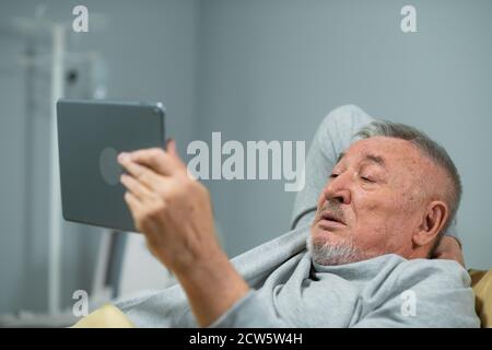 Älterer Mann, Patient, hält ein digitales Tablet in der Hand und schaut einen Film, während im Bett in einer Krankenstation bleiben, gesunde starke medizinische Konzept. Stockfoto