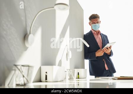 Porträt eines gutaussehenden Geschäftsmann trägt Maske und Blick auf die Kamera, während an der Wand lehnen in minimalem weißen Büro, kopieren Raum Stockfoto