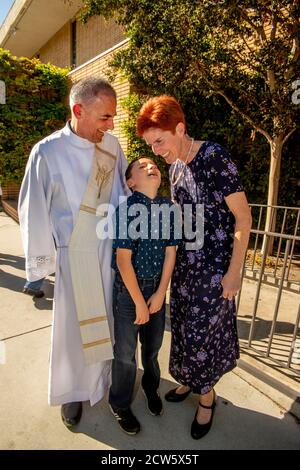 Nach der Messe posiert der Diakon einer katholischen Kirche in Südkalifornien mit einer Pfarrerin und ihrem Sohn im Kirchenaußenhof. Stockfoto