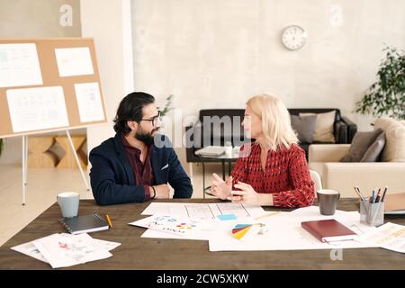 Junge bärtige Designerin und seine glückliche matue blonde Kollegin Sprechen Stockfoto