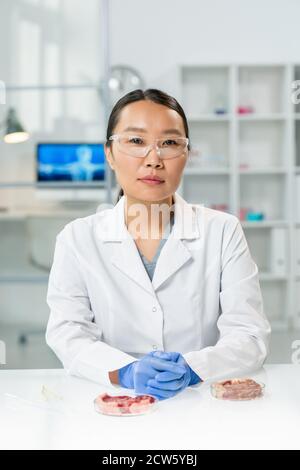 Junge, von jungen Menschen geliebte asiatische Forscherin in Wildlecoat und Brillen sitzen vorbei Arbeitsplatz Stockfoto