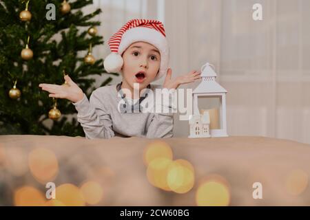 Überraschender Junge, in gestreifter Mütze und Fliege, mit offenen Augen, offenem Mund und verdünnten Händen. In der Neujahrsstimmung. Stockfoto