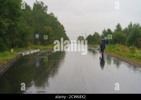 Der Herbst kommt in die russische Landschaft. Nasse Straße tagsüber. Kalte Dusche. Einsame Frau zog Regenmantel an, um zur Bushaltestelle zu laufen. Inlandstourismus Stockfoto