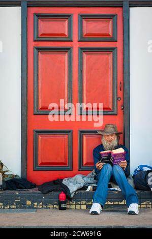 Ein alter Mann sitzt vor einer rot-schwarzen Tür und liest einen Roman, während er eine Pause von seiner Solo-Reise durch Townsville in QLD, Australien macht. Stockfoto