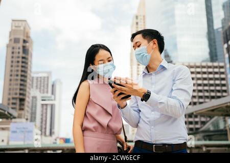 Asiatische Geschäftsmann und Frau tragen Covid Gesichtsbedeckungen Blick auf Handy in städtischen Umgebung, Stadtleben, Pandemie, Arbeitsleben, Kollegen Stockfoto