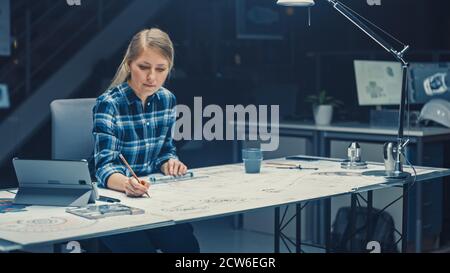 Weibliche Ingenieurin sitzt an ihrem Schreibtisch arbeitet mit Blueprints Laying on a Table, verwendet Bleistift, Lineal und Digital Tablet. Im dunklen Industriedesign Stockfoto