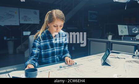 Weibliche Ingenieurin sitzt an ihrem Schreibtisch arbeitet mit Blueprints Laying on a Table, verwendet Bleistift, Lineal und Digital Tablet und trinkt Kaffee. Im Dunkeln Stockfoto