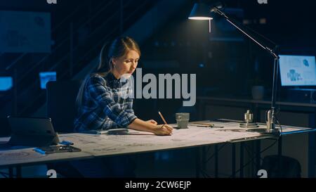 Weibliche Ingenieurin sitzt an ihrem Schreibtisch arbeitet mit Blueprints Laying on a Table, verwendet Bleistift, Lineal und Digital Tablet. Im dunklen Industriedesign Stockfoto