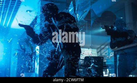 Maskierte Truppe bewaffneter SWAT-Polizisten stürmt ein finsteres beschlagnahmtes Bürogebäude mit Schreibtischen und Computern. Soldat bricht ein Glas mit seinem Arm und seinem Team Stockfoto