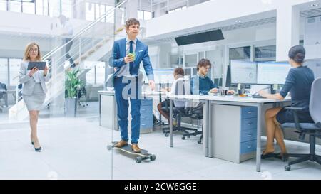 Im hellen Corporate Office fährt Cool Geschäftsmann Skateboard durch den Büroflur, hält Tasse mit Kaffee. Im Hintergrund Professional People Stockfoto