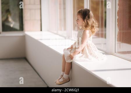 Nettes kleines Mädchen drei Jahre alt im Kleid posiert für Photograper im Studio. Wunderschönes kleines Mädchen sitzt auf der Fensterbank Stockfoto