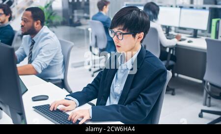 Junge professionelle japanische Aktienmakler arbeiten auf seinem Desktop-Computer. Vielfältige und motivierte Geschäftsleute arbeiten im modernen Open Office. Stockfoto