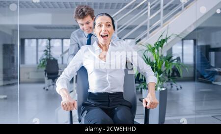 Fröhlicher Mitarbeiter schiebt seine schöne Kollegin auf einen Stuhl Zwischen Reihen von Schreibtischen, an denen verschiedene Geschäftsleute arbeiten Desktop-Computer in Stockfoto