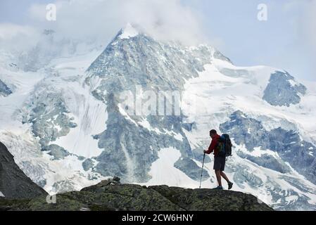 Männlicher Tourist mit Rucksack zu Fuß mit Trekkingstöcken. Erstaunlicher, felsiger Berg Ober Gabelhorn im Schnee in den Pennine Alpen in der Schweiz im Hintergrund. Konzept von Tourismus, Wandern, Alpinismus Stockfoto