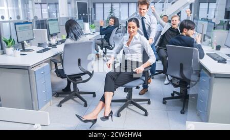Fröhlicher Mitarbeiter schiebt seine schöne Kollegin auf einen Stuhl Zwischen Reihen von Schreibtischen, an denen verschiedene Geschäftsleute arbeiten Desktop-Computer in Stockfoto