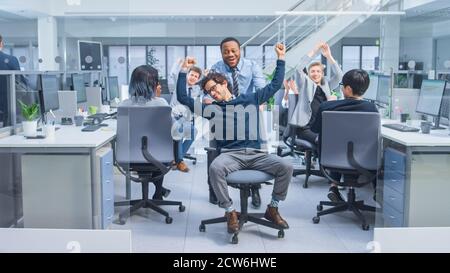 Fröhlicher Mitarbeiter schiebt seinen gutaussehenden Kollegen auf einen Stuhl dazwischen Reihen von Schreibtischen mit verschiedenen Geschäftsleuten, die am Desktop arbeiten Computer in modernen Stockfoto