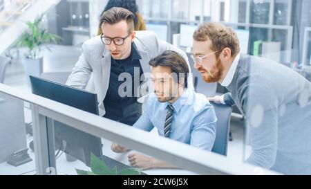 In Bright Modern Office: Junger Geschäftsmann sitzen und arbeiten an seinem Desktop-Computer mit Projektmanager und Teamleiter neben ihm stehen, haben Stockfoto