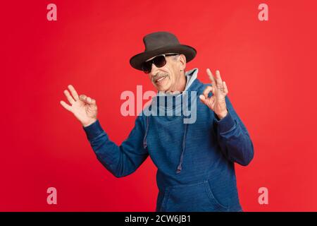 Schön, cool. Portrait von älteren Mann in stilvollen Brillen und Hut isoliert auf rotem Studio Hintergrund. Tech und fröhliche ältere Lifestyle-Konzept. Trendige Farben, für immer Jugend. Copyspace für Ihre Anzeige. Stockfoto