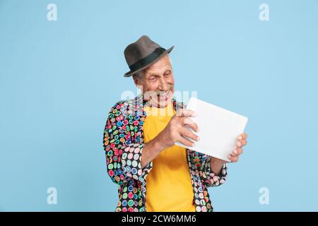 Spiele mit Tablet spielen. Portrait von Senior Hipster Mann in stilvollen Hut isoliert auf blauem Studio Hintergrund. Tech und fröhliche ältere Lifestyle-Konzept. Trendige Farben, für immer Jugend. Copyspace für Werbung. Stockfoto