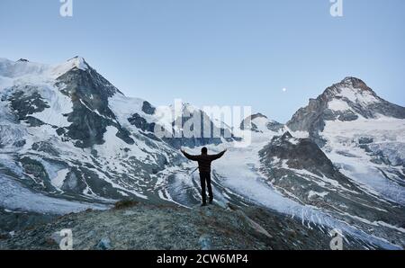 Fröhlicher Reisender, der die Nacht in den Bergen begrüßt, mit dem Rücken zur Kamera mit offenen Händen auf Gletscher und hohe schneebedeckte Gipfel der Pennine Alpen - Ober Gabelhorn und Dent Blanche schaut Stockfoto
