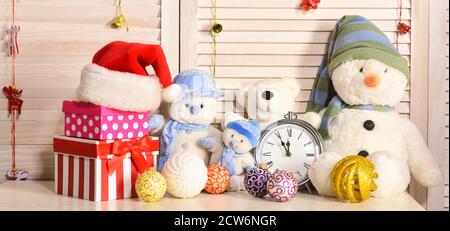 Spielzeug auf Büro auf Holzwand Hintergrund platziert. Weihnachtsdekoration im festlichen Raum. Schneemänner, Teddybären, Weihnachtskugeln und Geschenkboxen in der Nähe des Weckers. Feier- und Neujahrsdekor-Konzept Stockfoto