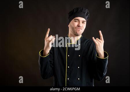 Porträt eines jungen Mannes in einem schwarzen Koch Anzug gekleidet Tun Rock-Symbol mit Händen oben posieren auf einem braunen Hintergrund mit Kopierraum Werbefläche Stockfoto