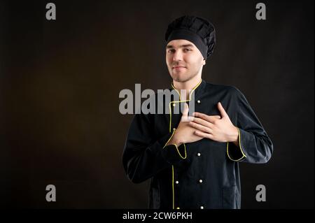 Porträt eines jungen Mannes in einem schwarzen Koch Anzug gekleidet Hält seine Hände auf der Brust neben seinem Herzen posiert Auf braunem Hintergrund mit Kopierbereich Stockfoto