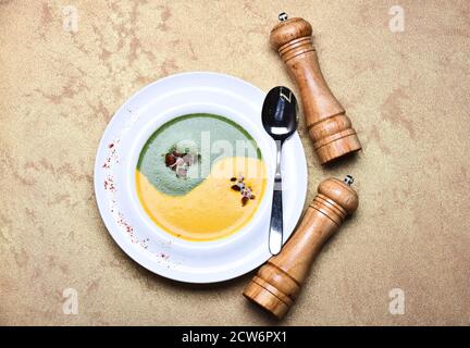 Traditionelles Küchenkonzept. Japanische Suppe im Restaurant. Püree in grüner und gelber Farbe in Yin Yang Zeichen durch Pfefferbox, Salzkeller und Löffel. Spinat- und Kürbiscremesuppe auf grauem Texturhintergrund Stockfoto