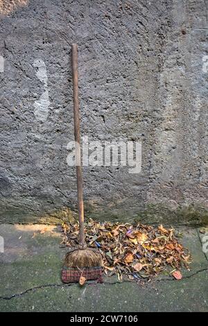 An die Wand gelehnt steht ein abgenutzter alter Besen und heruntergefallene Blätter auf einem Haufen. Stockfoto