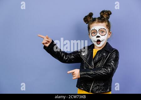 Portrait von niedlichen kleinen Mädchen mit hellen gruseligen Make-up, in schwarzer Lederjacke gekleidet, zeigt neben auf lila Studio kopieren Raum, zeigt Ort von Hallo Stockfoto