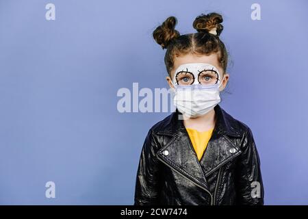 Portrait of Little girl hat funky Make-up und schützende medizinische Maske, in schwarzer Kleidung gekleidet, isoliert auf lila Studio-Hintergrund mit Kopieplatz f Stockfoto