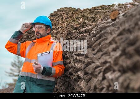 Forsttechniker, der mit einem Notizblock in der Zwischenablage neben einem Baumlogan in der Walde posiert, ist zuversichtlich, dass männliche Fachmann Daten auf dem Feld sammelt Stockfoto