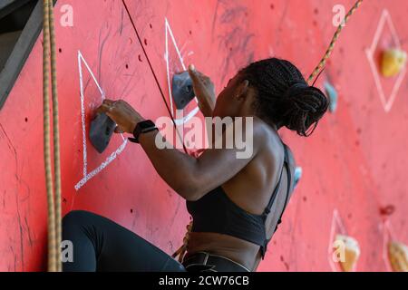 Junge schwarze Frau klettert auf Trainingskletterwand. Afrikanisches Klettermädchen geht durch die Route Stockfoto