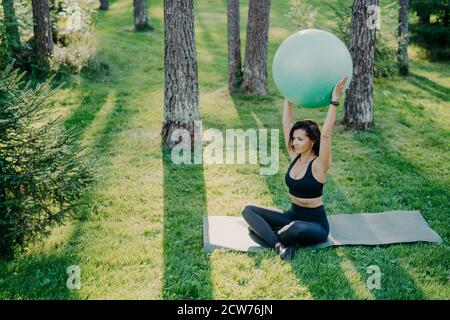 Fit sportliche Frau Übungen mit Fitness-Ball sitzt auf Karemat in Lotushaltung gekleidet in Cropped Top und Leggings, Posen im Wald während sonnigen Tag, Brea Stockfoto