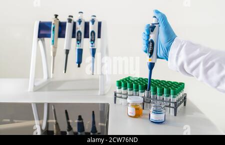 Pipettierassistent mit einer Mikropipette auf weißem Hintergrund. Hand in blauen Handschuh tropfende Flüssigkeit auf Reagenzglas und verschiedene Pipetten in Kunststoff-Rack. Stockfoto