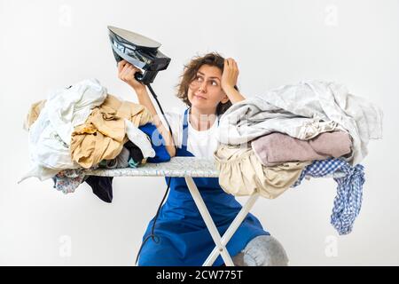 Eine Frau hält ein Bügeleisen in den Händen und sitzt am Bügeltisch. Schmutzige Wäsche vor dem Bügeln nach dem Waschen. Reinigung und Waschen und Bügeln Konzept Stockfoto