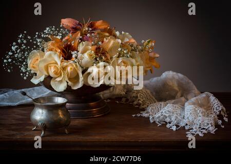 Stillleben Bouquet mit cremigen weißen und orangen Rosen und Lilien in einer Zinnschale mit einem Spitzenschal Stockfoto