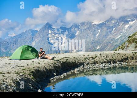 Männlicher Tourist sitzt im Stuhl neben dem Zelt in den felsigen alpinen Bergen nahe dem See mit frischem klarem Wasser und mit dem Smartphone an sonnigen warmen Sommertagen. Konzept von Camping, Reisen, Natur, Gadgets. Stockfoto