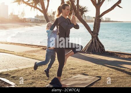 Junge Mutter und Sohn machen Gymnastik und Stretching im Stadtpark auf Sonnenuntergang mit Meerblick. Frau im Yoga Asana und Junge auf ihren Schultern Stockfoto