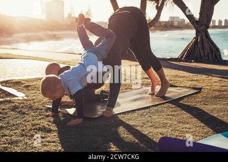 Junge Mutter und zwei Söhne machen Gymnastik und Stretching im Stadtpark bei Sonnenuntergang mit Meerblick. Frau in Yoga Asana und Junge auf dem Rücken Stockfoto