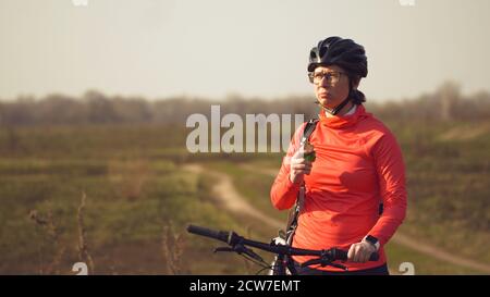 Athletische kaukasische Frau isst Protein-Bar-Fahrt auf dem Mountainbike auf der Natur. Junge sportliche Frau Athlet in Helm ruhen während Beißen Ernährungsriegel Stockfoto