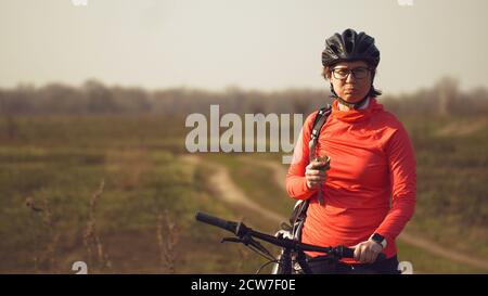 Athletische kaukasische Frau isst Protein-Bar-Fahrt auf dem Mountainbike auf der Natur. Junge sportliche Frau Athlet in Helm ruhen während Beißen Ernährungsriegel Stockfoto