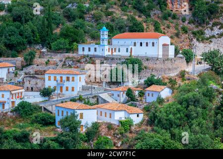 Blick auf das albanische Dorf - traditionelle weiße Häuser mit orangefarbenen Dächern und hölzernen Fensterläden - auf den Berg, kleine Kirche und Straße. Stockfoto