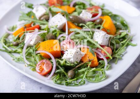 Warmer Salat mit Kürbis, Feta-Käse, Tomaten, Kapern, Rucola und roten Zwiebeln. Nahaufnahme. Stockfoto