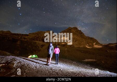 Rückansicht der Wanderer bewundern Blick auf majestätische Berge unter Nachthimmel mit Starts. Frau und Mann stehen auf einem felsigen Hügel und genießen die nächtliche Berglandschaft. Konzept von Reisen, Wandern, Nacht. Stockfoto