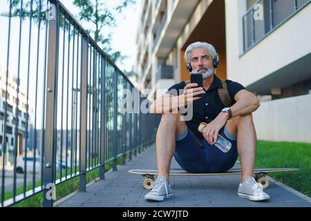 Porträt eines reifen Mannes mit Kopfhörern, die im Freien in der Stadt sitzen, mit dem Smartphone. Stockfoto