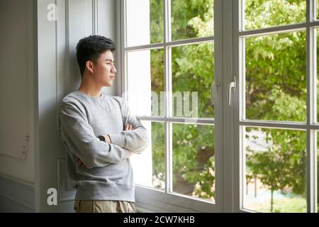 Junger asiatischer Mann steht am Fenster zu Hause Arme gekreuzt Stockfoto