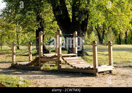 Hölzerne Spielbrücke für Kinder, im Park unter den Bäumen gelegen. Spielmöglichkeiten im Freien. Brücke aus Holzbohlen mit Seilgeländer. Stockfoto